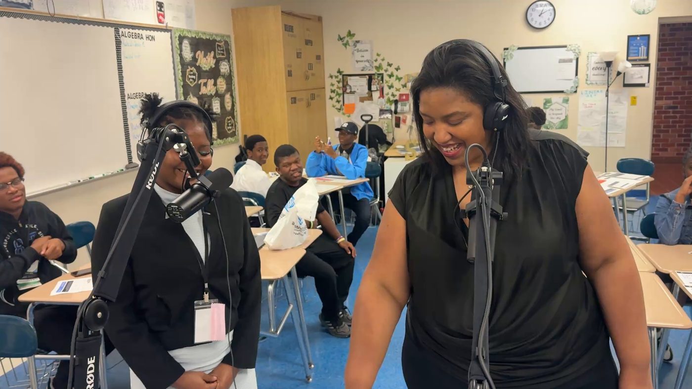 A facilitator in a black top and headphones laughs at a microphone while a student in headphones stands beside her. Other students sit at desks behind them.