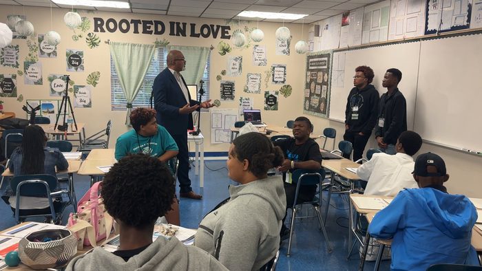 A man in a navy blazer talks with a classroom of students under a Rooted in Love banner while two students stand at the whiteboard.