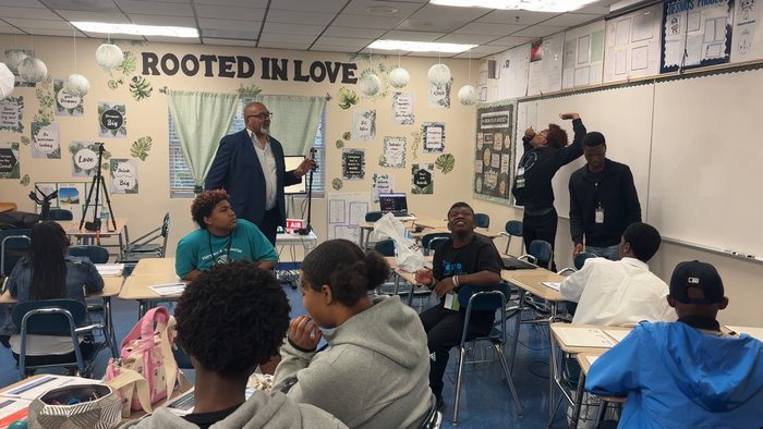 Two students write on the whiteboard while the man in the navy blazer watches and the class looks on.
