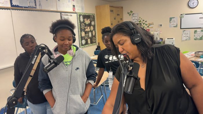 A student in a gray hoodie and headphones stands at a microphone beside the facilitator, with other teens behind them.
