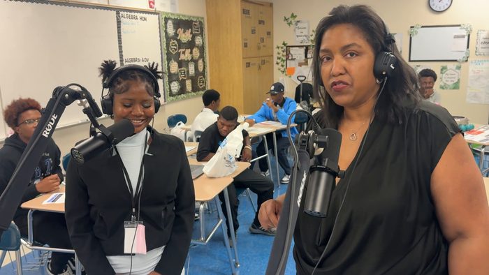 A student at a microphone and the facilitator in headphones, with classmates seated at desks in the background.