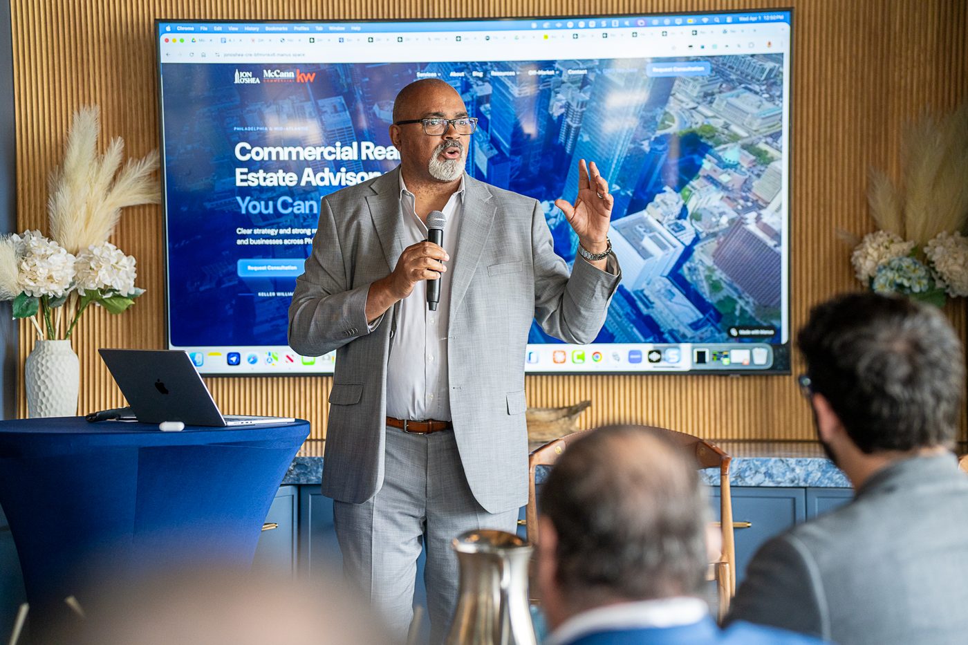 Ross Jones, a bald man with glasses and a gray beard, in a light gray blazer, speaks into a handheld microphone and gestures with one hand. A large screen behind him shows a commercial real estate website.