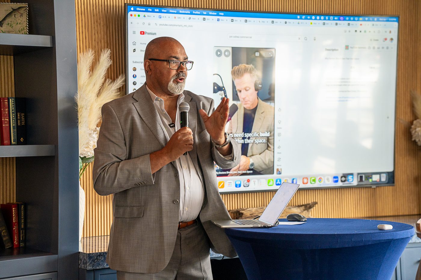 Ross Jones in a gray blazer speaks into a handheld microphone with his hand raised, standing beside a laptop on a navy-draped table. A large screen behind him shows a short video.