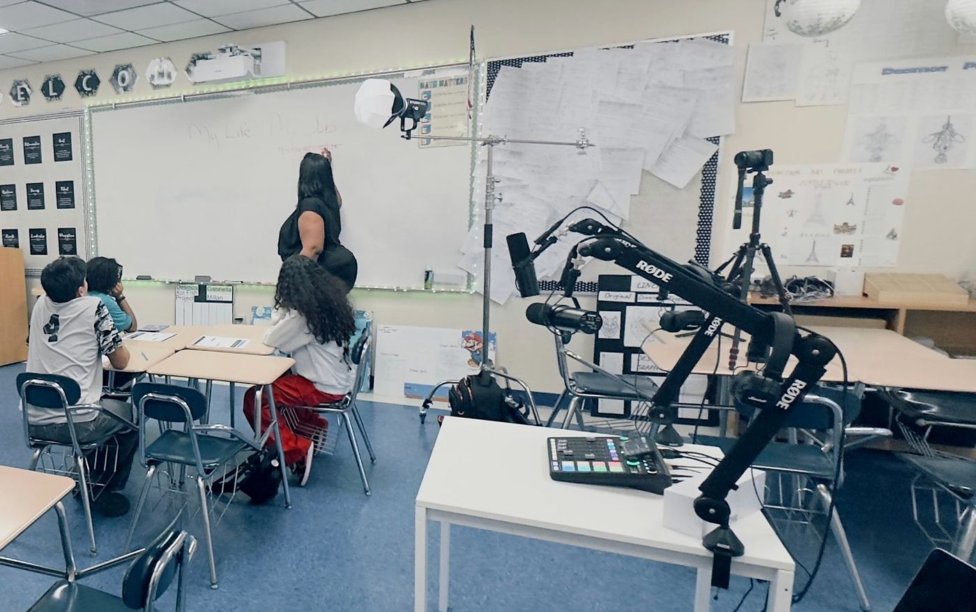 Students sit at desks facing a whiteboard where a facilitator is writing. A boom-arm microphone, a mixing board and a camera on a tripod are set up beside the whiteboard.