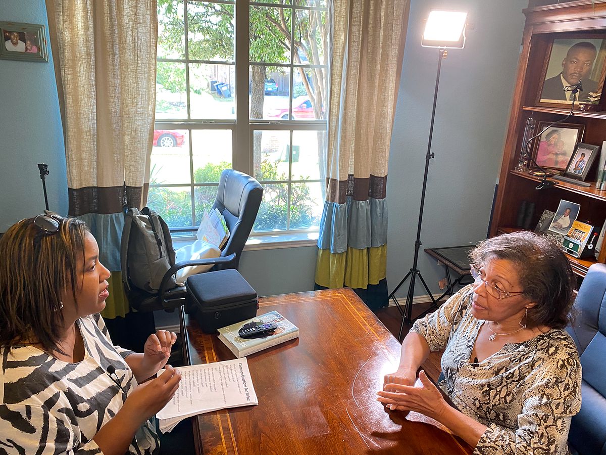 Two women sit across a wooden desk in a home office. One holds notes and wears a small clip-on microphone while the other talks with her hands. A studio light stands behind them.