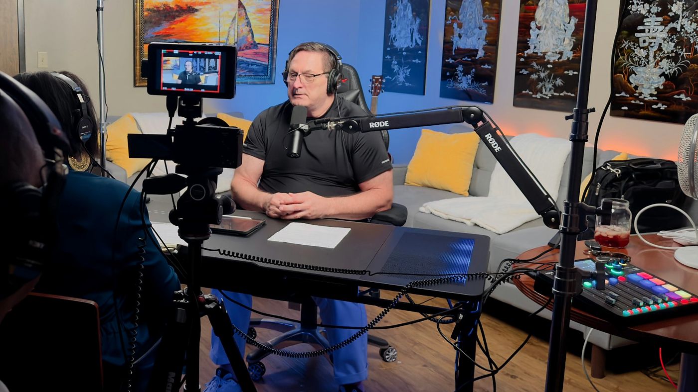 A man in a black shirt speaks at a studio microphone on a boom arm. A camera on a tripod, studio lights and colorful artwork fill the room.